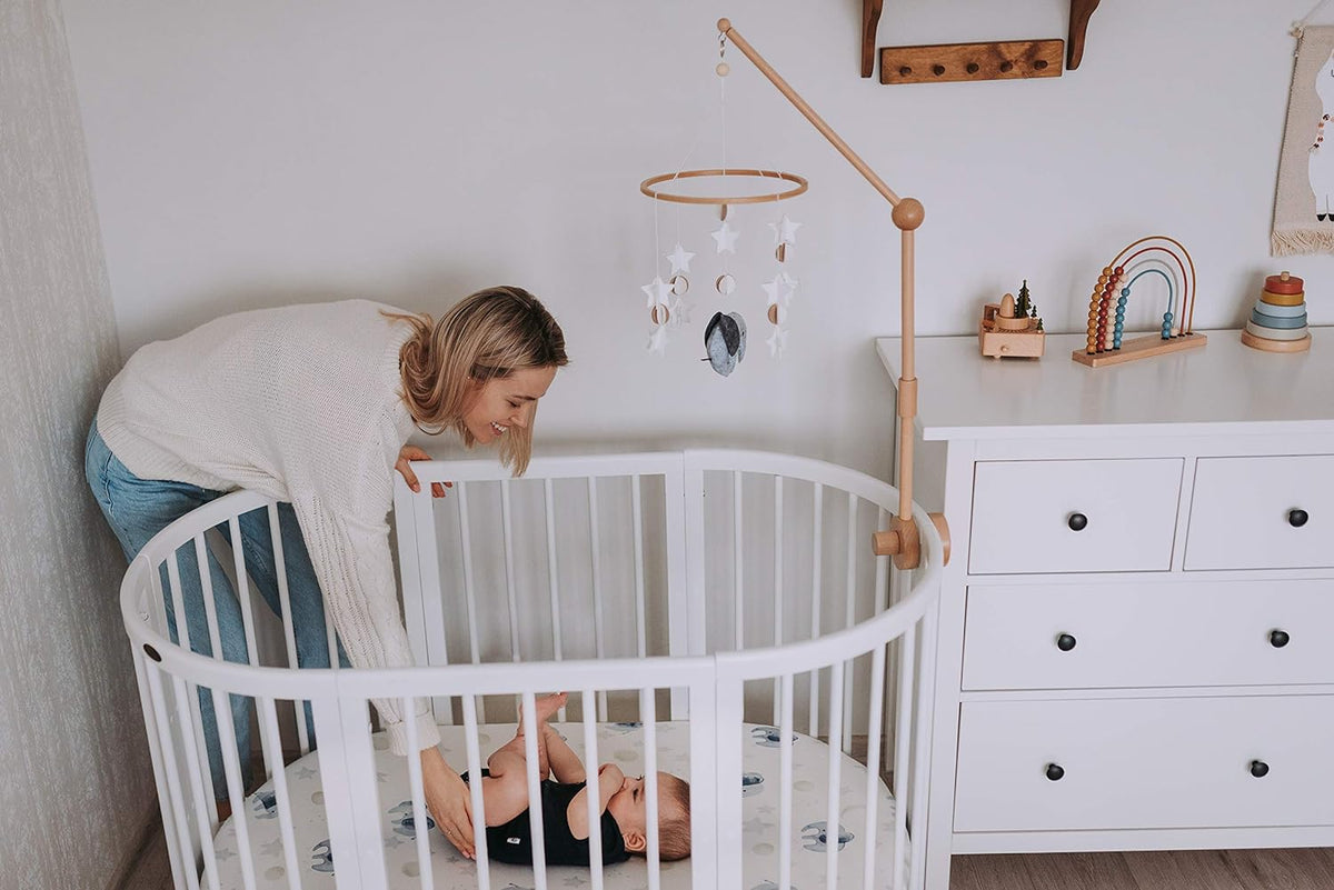 Mother leaning over a white oval crib to her baby, with a Moon and Stars mobile above