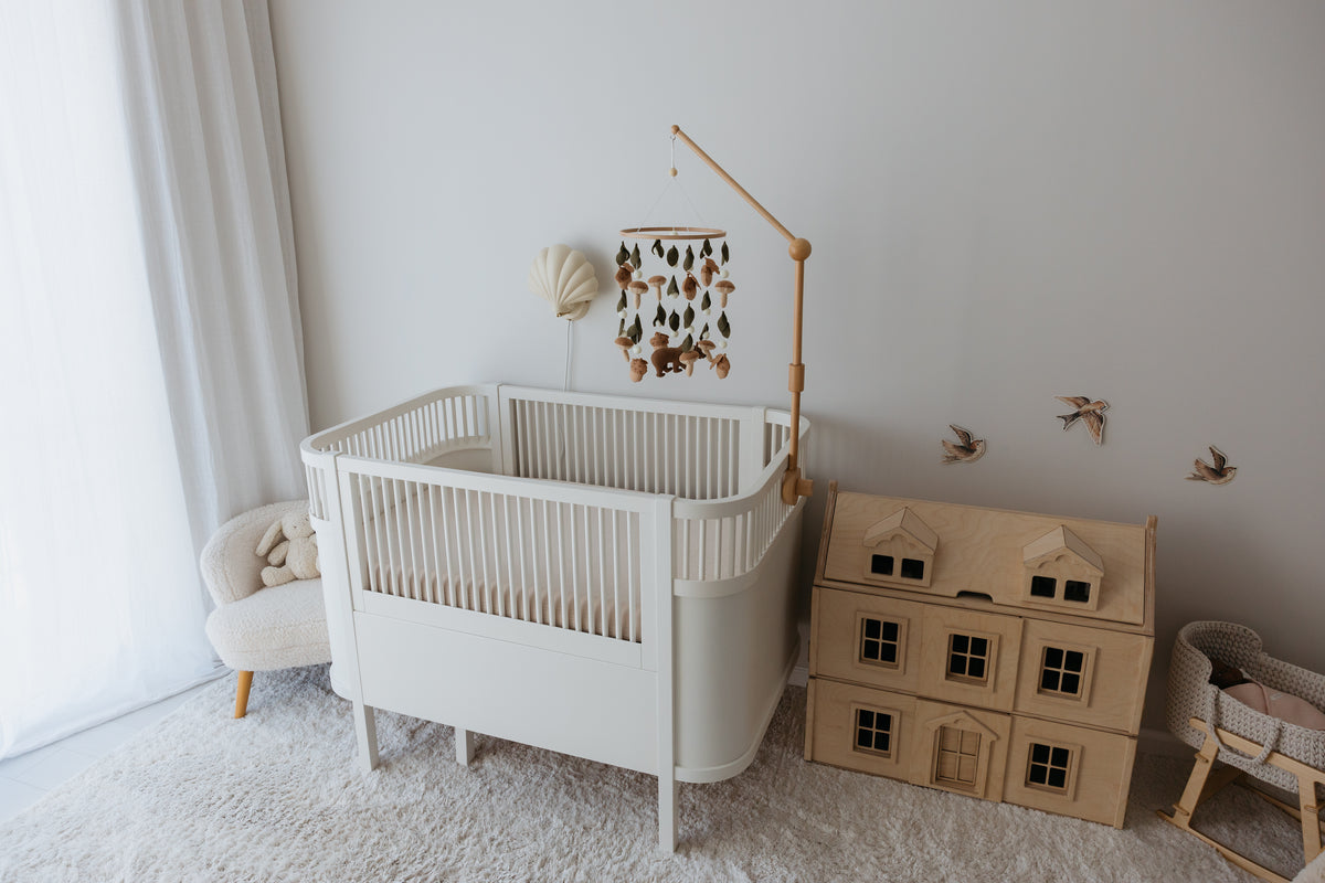 White crib with a Magic Forest mobile beside a wooden dollhouse in a calm nursery